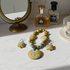 A handmade beaded necklace and matching drop earrings with a colorful floral pattern, styled on a marble surface with perfume bottles and a mirror in the background.