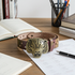 A leather belt with a large bronze tiger head buckle, displayed on a wooden desk surrounded by books, papers, and a pen.