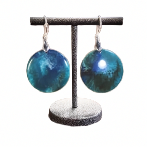 A pair of round drop earrings with a marbled blue and teal pattern, hanging from silver hooks on a black jewelry display stand against a white background.