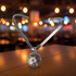A polished silver metal heart-shaped necklace with a circular pendant, displayed on a wooden table with a warm, blurred bokeh background of lights.