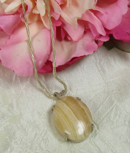 A silver-toned necklace with an oval pendant made from a polished, striped natural shell, resting on a textured white fabric next to pink flowers.