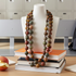 A long beaded necklace made of round earthy-toned beads draped over a white jewelry bust, styled on top of stacked books on a desk with a blurred classroom or office background.