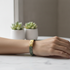 A close-up of a person's wrist wearing a gold-tone rectangular bracelet with a dark green and white patterned band, resting on a white marble surface with small potted succulents in the background.
