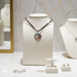 A silver-toned necklace with a round pendant featuring a stylized eye design, displayed on a white jewelry bust. The bust is set on a marble surface with other jewelry pieces, including a bracelet, earrings, and another necklace, arranged nearby in a boutique display.
